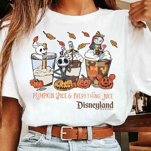 May include: White t-shirt featuring a colorful graphic of three themed beverages with Jack Skellington, Zero, and Sally from The Nightmare Before Christmas. The text reads "Pumpkin Spice & Everything Nice Disneyland Resort."