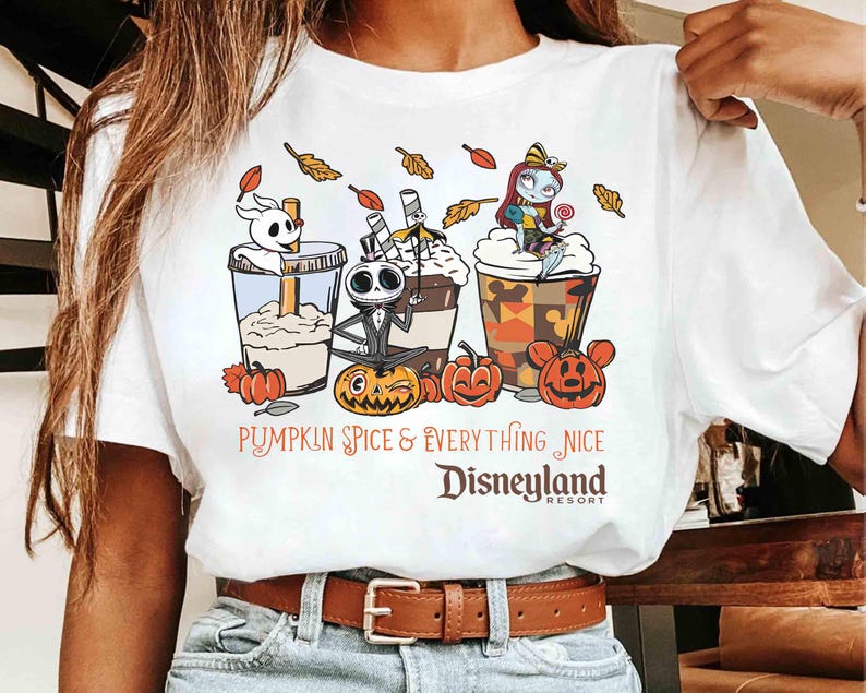 May include: White t-shirt featuring a colorful graphic of three themed beverages with Jack Skellington, Zero, and Sally from The Nightmare Before Christmas. The text reads "Pumpkin Spice & Everything Nice Disneyland Resort."
