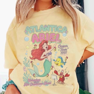 May include: A pale yellow t-shirt featuring a graphic print of Ariel the mermaid, Flounder, and Sebastian. The text reads ATLANTICA ARIEL Ocean Tour 1991 and Sebastian with the Hot Crustacean Band, surrounded by colorful coral and bubbles.