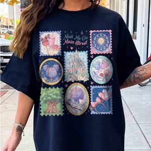 Peut inclure: Un t-shirt bleu marine avec un graphisme color&eacute; d'illustrations de style timbre-poste. Les timbres repr&eacute;sentent diverses ic&ocirc;nes de parc &agrave; th&egrave;me, dont un ch&acirc;teau, une grande roue et un arbre. Le texte "Meet Me On Main Street" est imprim&eacute; au-dessus des timbres.