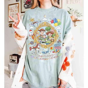May include: A light blue t-shirt featuring a colorful graphic of characters from the animated movie Toy Story. The design includes Woody, Buzz Lightyear, and other characters within a gold frame, with the text "YOU'VE GOT A FRIEND IN ME".