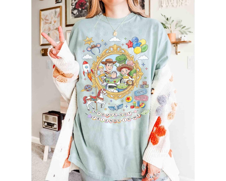 May include: A light blue t-shirt featuring a colorful graphic of characters from the animated movie Toy Story. The design includes Woody, Buzz Lightyear, and other characters within a gold frame, with the text "YOU'VE GOT A FRIEND IN ME".