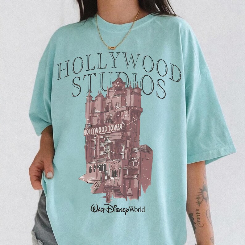 May include: A light blue t-shirt featuring a graphic of the Hollywood Tower Hotel. The text reads HOLLYWOOD STUDIOS at the top, The HOLLYWOOD TOWER on the building, and Walt Disney World at the bottom. The design is printed in shades of brown.