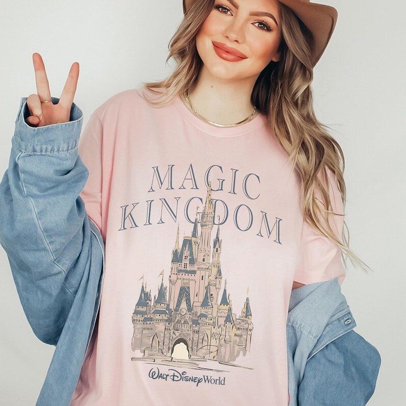 May include: A light pink short-sleeved t-shirt featuring the text MAGIC KINGDOM in dark blue lettering above a detailed illustration of a castle and the Walt Disney World logo. The shirt is worn under a light blue denim button-down shirt.
