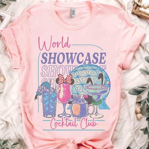 以下が含まれることがあります： 「World Showcase Cocktail Club」の文字とカクテルと幾何学的な球体のグラフィックがプリントされたピンクのTシャツ。Tシャツは袖がロールアップされ、クルーネックです。