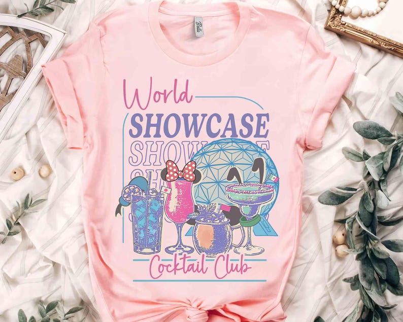 以下が含まれることがあります： 「World Showcase Cocktail Club」の文字とカクテルと幾何学的な球体のグラフィックがプリントされたピンクのTシャツ。Tシャツは袖がロールアップされ、クルーネックです。