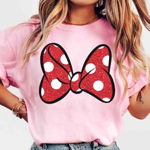 Puede incluir: Camiseta rosa claro con un gran lazo rojo con lunares blancos. El lazo tiene una textura brillante y un contorno negro. La camiseta es de color liso y tiene mangas cortas.