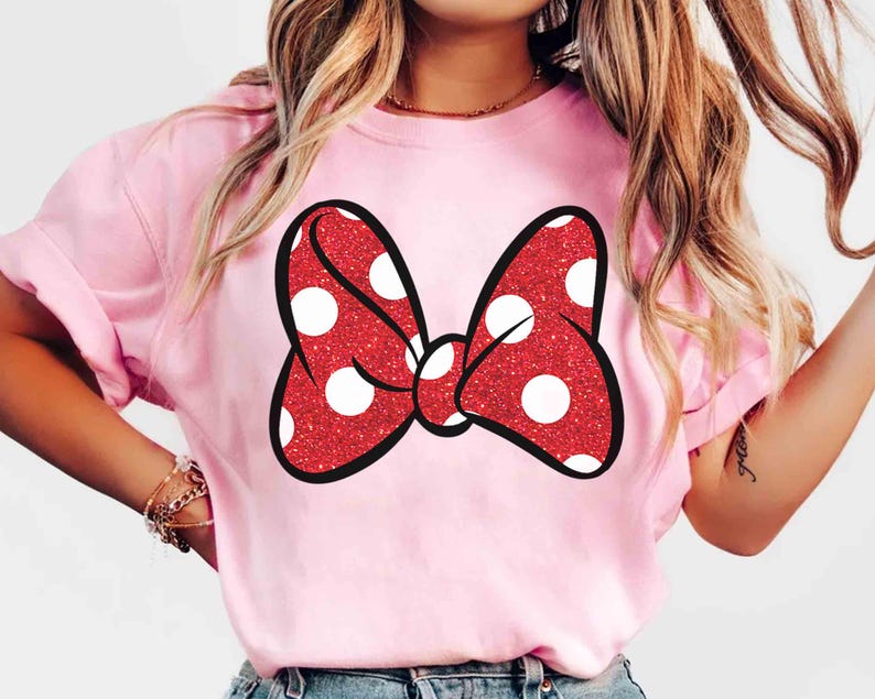 Puede incluir: Camiseta rosa claro con un gran lazo rojo con lunares blancos. El lazo tiene una textura brillante y un contorno negro. La camiseta es de color liso y tiene mangas cortas.