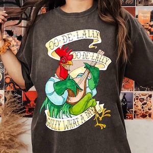 Op de afbeelding: Donkergrijs T-shirt met een cartoon haan die een luit bespeelt, met de tekst "OO-DE-LALLY" en "GOLLY WHAT A DAY" op banners. De haan is rood, groen en blauw. Het shirt is een casual stijl.