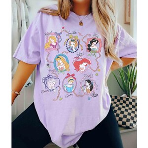 Puede incluir: Una camiseta de color lila claro con retratos enmarcados de princesas animadas. Cada retrato est&aacute; en un marco decorativo y conectado por una cadena. La camiseta tiene mangas cortas y un corte holgado.