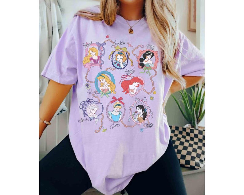 Puede incluir: Una camiseta de color lila claro con retratos enmarcados de princesas animadas. Cada retrato est&aacute; en un marco decorativo y conectado por una cadena. La camiseta tiene mangas cortas y un corte holgado.