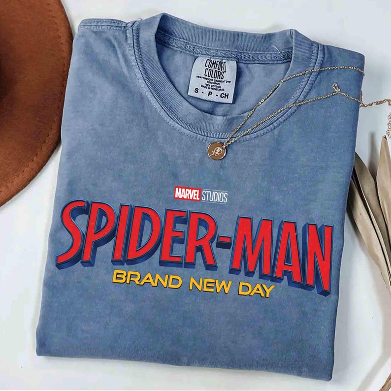 May include: A blue t-shirt featuring the Marvel Studios Spider-Man logo in red and blue with the words "BRAND NEW DAY" in yellow. A gold necklace with a pendant rests on the shirt. The shirt is folded on a white surface.