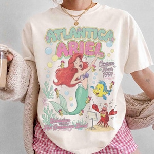 May include: A cream-colored t-shirt featuring a graphic print of Ariel the mermaid, Flounder, and Sebastian. The text reads Atlantica Ariel, Ocean Tour 1991, and Sebastian with the Hot Crustacean Band. The design includes colorful bubbles and coral.