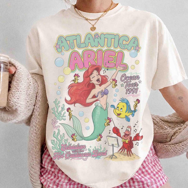 May include: A cream-colored t-shirt featuring a graphic print of Ariel the mermaid, Flounder, and Sebastian. The text reads Atlantica Ariel, Ocean Tour 1991, and Sebastian with the Hot Crustacean Band. The design includes colorful bubbles and coral.
