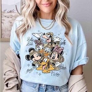 May include: Pale blue t-shirt with a graphic of Disney characters Mickey Mouse, Minnie Mouse, Donald Duck, Daisy Duck, Goofy, and Pluto. The characters are wearing safari hats. The shirt has the names of the characters and the year 2020.