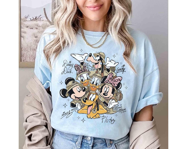 May include: Pale blue t-shirt with a graphic of Disney characters Mickey Mouse, Minnie Mouse, Donald Duck, Daisy Duck, Goofy, and Pluto. The characters are wearing safari hats. The shirt has the names of the characters and the year 2020.