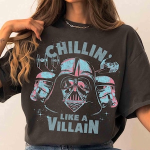 Può includere: T-shirt grigio scuro con grafica di Darth Vader e Stormtrooper in blu e rosa. Il testo "CHILLIN' LIKE A VILLAIN" è stampato sopra e sotto l'immagine in blu.