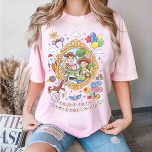 May include: A light pink t-shirt featuring a colorful graphic from the movie Toy Story. The design includes characters like Woody, Buzz Lightyear, and Rex, set within a gold frame. The text "YOU'VE GOT A FRIEND IN ME" is also included.