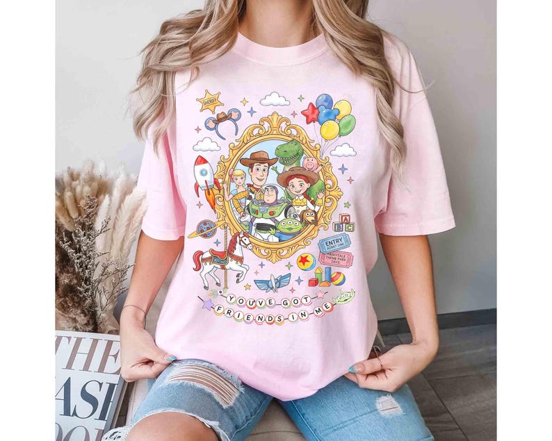 May include: A light pink t-shirt featuring a colorful graphic from the movie Toy Story. The design includes characters like Woody, Buzz Lightyear, and Rex, set within a gold frame. The text "YOU'VE GOT A FRIEND IN ME" is also included.