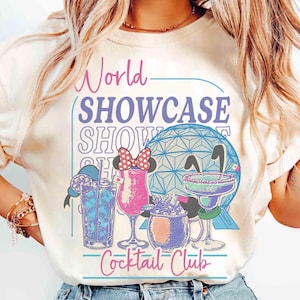 以下が含まれることがあります： 「World Showcase Cocktail Club」の文字とカクテルのイラストが描かれたクリーム色のTシャツ。幾何学的な球体と青、ピンク、紫の色使いが特徴で、楽しくカラフルなグラフィックです。