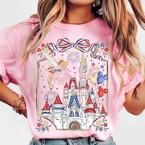 Puede incluir: Camiseta rosa claro con un gr&aacute;fico colorido de un hada, fuegos artificiales, un castillo y globos de Mickey Mouse. El dise&ntilde;o incluye elementos rojos, blancos y azules, y un lazo decorativo. Es una prenda casual.