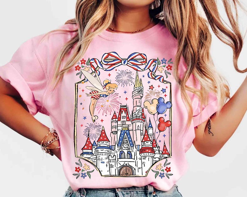 Puede incluir: Camiseta rosa claro con un gr&aacute;fico colorido de un hada, fuegos artificiales, un castillo y globos de Mickey Mouse. El dise&ntilde;o incluye elementos rojos, blancos y azules, y un lazo decorativo. Es una prenda casual.