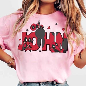 May include: Pale pink t-shirt with the name "JOHN" in red, accompanied by Deadpool character illustrations. The design incorporates Deadpool figures in different poses and red star motifs. The t-shirt is a light pink colour.