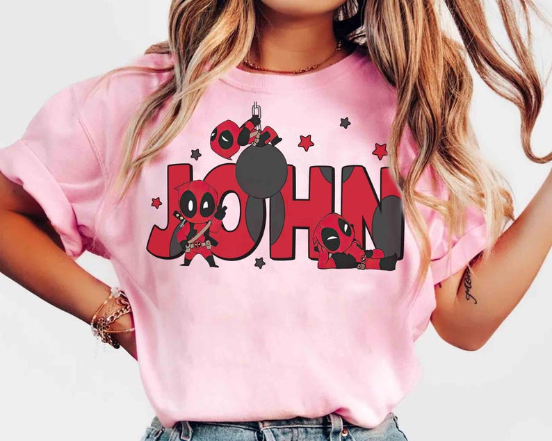 May include: Pale pink t-shirt with the name "JOHN" in red, accompanied by Deadpool character illustrations. The design incorporates Deadpool figures in different poses and red star motifs. The t-shirt is a light pink colour.
