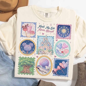 Peut inclure: T-shirt cr&egrave;me avec un graphisme color&eacute; d'illustrations de style timbre-poste. Les timbres repr&eacute;sentent diverses ic&ocirc;nes de parc &agrave; th&egrave;me, dont un ch&acirc;teau, une grande roue et des friandises sur le th&egrave;me de Mickey Mouse. Le texte "Meet Me On Main Street" est &eacute;galement visible.