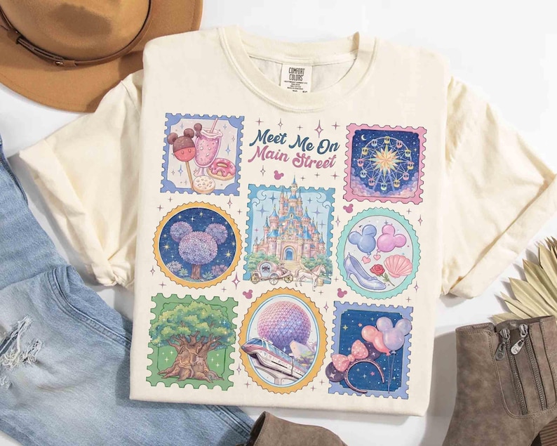 Peut inclure: T-shirt cr&egrave;me avec un graphisme color&eacute; d'illustrations de style timbre-poste. Les timbres repr&eacute;sentent diverses ic&ocirc;nes de parc &agrave; th&egrave;me, dont un ch&acirc;teau, une grande roue et des friandises sur le th&egrave;me de Mickey Mouse. Le texte "Meet Me On Main Street" est &eacute;galement visible.