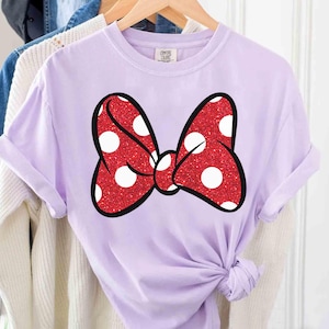 Puede incluir: Una camiseta lila con un gran lazo rojo con lunares blancos. El lazo tiene una textura brillante y un contorno negro. La camiseta est&aacute; colgada en una percha de madera con otras prendas en el fondo.