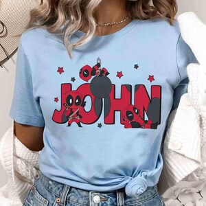 May include: Pale blue t-shirt with the name "JOHN" in red, accompanied by Deadpool character illustrations. The design includes Deadpool figures and red stars. The shirt is a short-sleeved crew neck.