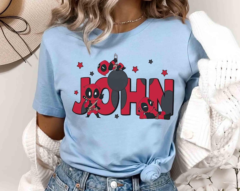 May include: Pale blue t-shirt with the name "JOHN" in red, accompanied by Deadpool character illustrations. The design includes Deadpool figures and red stars. The shirt is a short-sleeved crew neck.