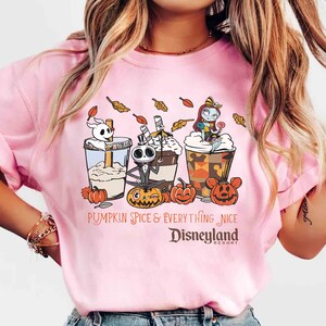 May include: Pink t-shirt featuring a graphic of three themed coffee drinks with characters and pumpkins. The text reads "Pumpkin Spice & Everything Nice Disneyland Resort". The design includes cartoon characters and fall leaves.