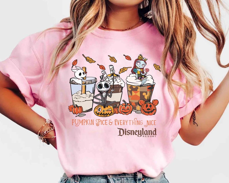 May include: Pink t-shirt featuring a graphic of three themed coffee drinks with characters and pumpkins. The text reads "Pumpkin Spice & Everything Nice Disneyland Resort". The design includes cartoon characters and fall leaves.