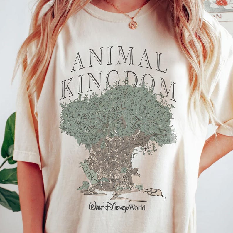 May include: A cream-colored short-sleeve t-shirt featuring the text ANIMAL KINGDOM in black capital letters above a graphic illustration of a large tree with green foliage and a brown trunk, with the text Walt Disney World printed below.