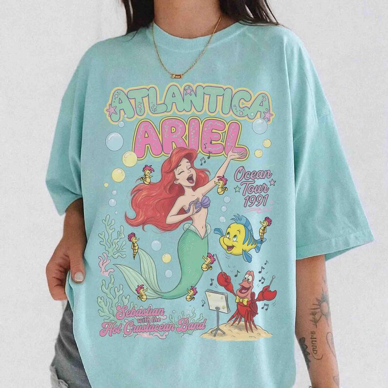 May include: A light blue t-shirt featuring a graphic print of Ariel from The Little Mermaid with the text Atlantica Ariel Ocean Tour 1991 and Sebastian with the Hot Crustacean Band. The design includes colorful illustrations of sea life.