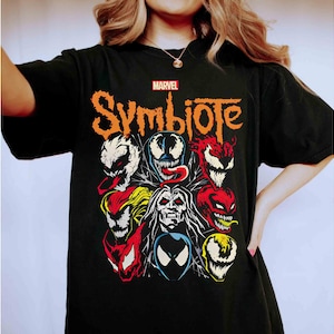 May include: Black t-shirt featuring a graphic of Marvel's Symbiote characters. The design includes various faces in red, white, yellow, and blue, with the word "Symbiote" in orange. The Marvel logo is at the top.