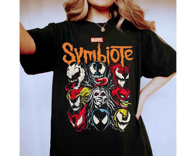 May include: Black t-shirt featuring a graphic of Marvel's Symbiote characters. The design includes various faces in red, white, yellow, and blue, with the word "Symbiote" in orange. The Marvel logo is at the top.