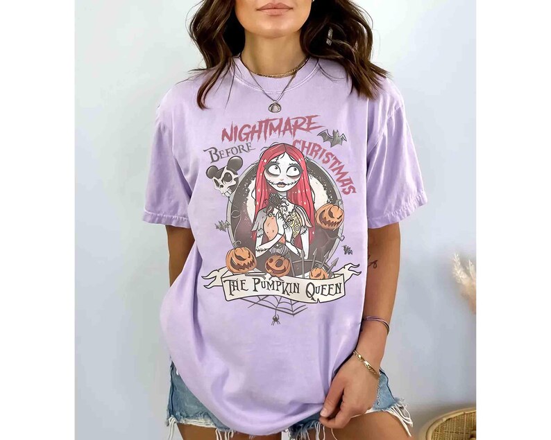 Puede incluir: Camiseta lila claro con un dise&ntilde;o gr&aacute;fico de "Pesadilla antes de Navidad". El dise&ntilde;o incluye a Sally, calabazas y el texto "The Pumpkin Queen". La camiseta tiene mangas cortas y cuello redondo.