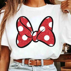 Puede incluir: Camiseta blanca con un gran lazo rojo con lunares blancos. El lazo tiene una textura brillante y un contorno negro. La camiseta se combina con vaqueros azules y un cintur&oacute;n marr&oacute;n.