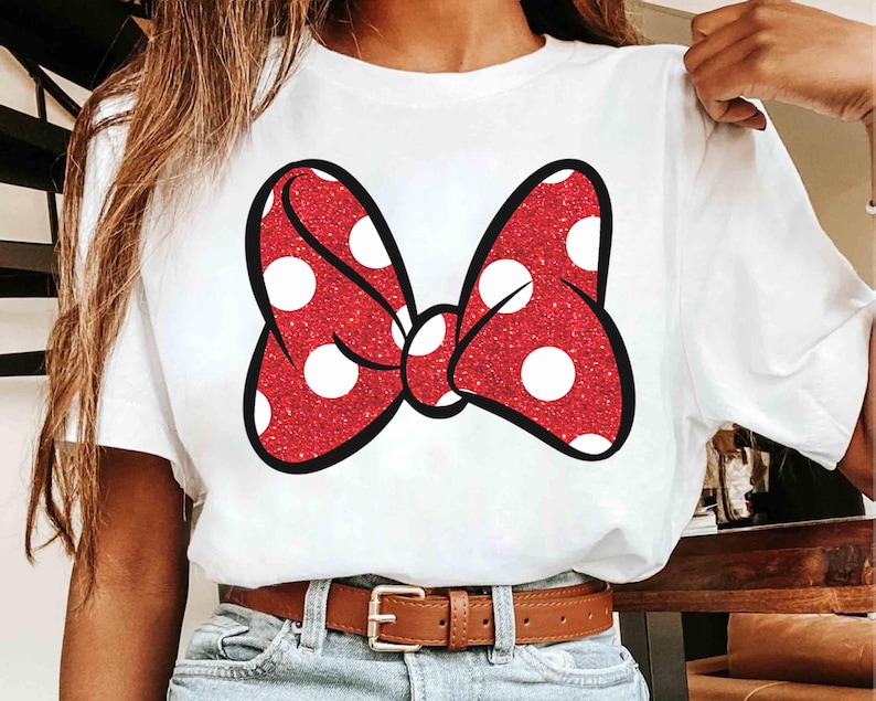 Puede incluir: Camiseta blanca con un gran lazo rojo con lunares blancos. El lazo tiene una textura brillante y un contorno negro. La camiseta se combina con vaqueros azules y un cintur&oacute;n marr&oacute;n.