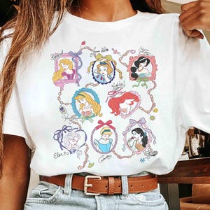 Puede incluir: Camiseta blanca con un gr&aacute;fico colorido de varias princesas de dibujos animados en marcos ornamentados. El dise&ntilde;o incluye los nombres de las princesas en escritura cursiva, conectados por una cadena.