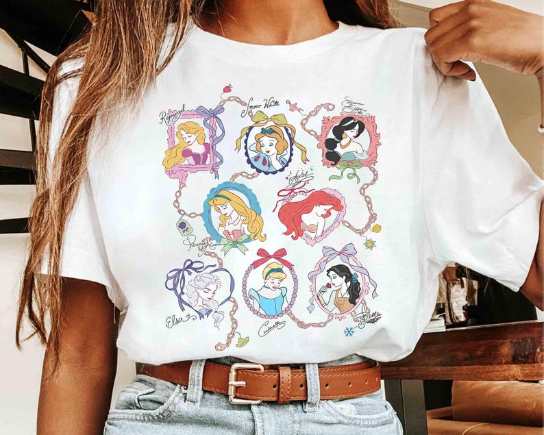 Puede incluir: Camiseta blanca con un gr&aacute;fico colorido de varias princesas de dibujos animados en marcos ornamentados. El dise&ntilde;o incluye los nombres de las princesas en escritura cursiva, conectados por una cadena.