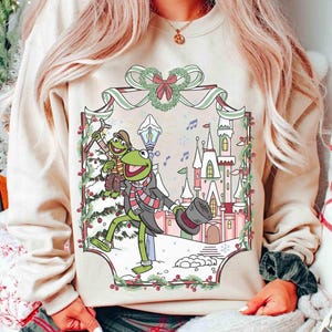 Puede incluir: Sudadera beige con una ilustración festiva de la Rana Gustavo en una escena invernal. El diseño incluye un castillo, nieve y notas musicales, con Gustavo usando un sombrero de copa y bufanda. Una corona y guirnalda navideñas enmarcan la imagen.