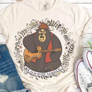 Könnte beinhalten: Cremefarbenes T-Shirt mit einem Cartoon-Bären, der Gitarre spielt, umgeben von einem floralen Kranz. Der Bär trägt einen Hut und eine Weste, die Gitarre ist mit "Big Al" beschriftet. Der Text "Blood on the Saddle" steht unter dem Bären.
