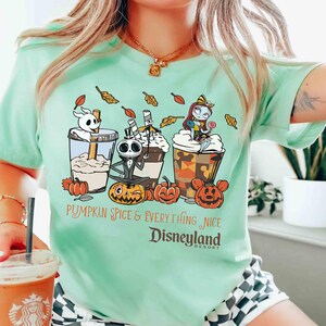 May include: A light green t-shirt featuring a graphic of three themed coffee drinks with Jack Skellington, Zero, and Sally from The Nightmare Before Christmas. The text reads "Pumpkin Spice & Everything Nice Disneyland Resort."