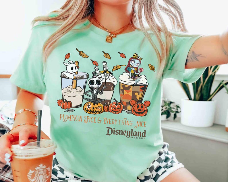 May include: A light green t-shirt featuring a graphic of three themed coffee drinks with Jack Skellington, Zero, and Sally from The Nightmare Before Christmas. The text reads "Pumpkin Spice & Everything Nice Disneyland Resort."