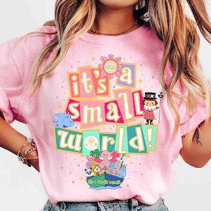 Op de afbeelding: Roze T-shirt met de tekst "it's a small world!" in kleurrijke blokletters. Het ontwerp bevat een bloem met het woord "hello", een stripfiguur en diverse grillige wezens in een boot. Het shirt heeft een ronde hals.