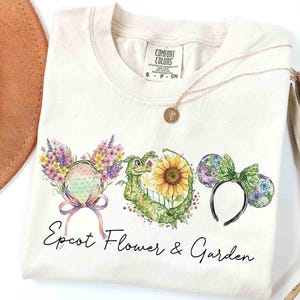 May include: Cream-colored t-shirt featuring a floral design with the text "Epcot Flower & Garden". The design includes colorful floral Mickey Mouse ears, a dragon, and a sunflower. A gold necklace is draped on the shirt.